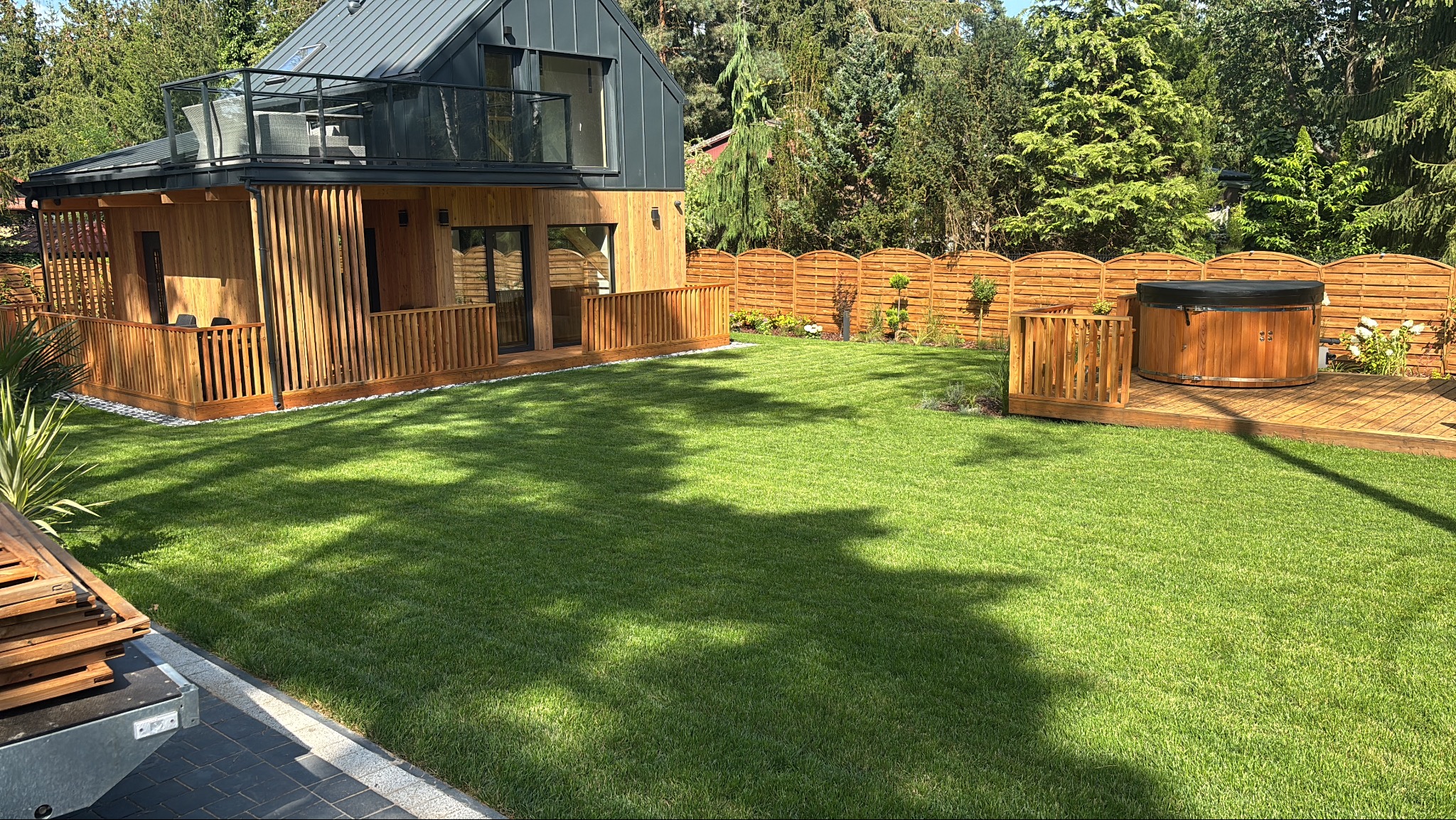 Nowoczesny dom z drewnianą elewacją, tarasem i jacuzzi na zielonym trawniku w słoneczny dzień. Minimalistyczny design z elementami naturalnymi.