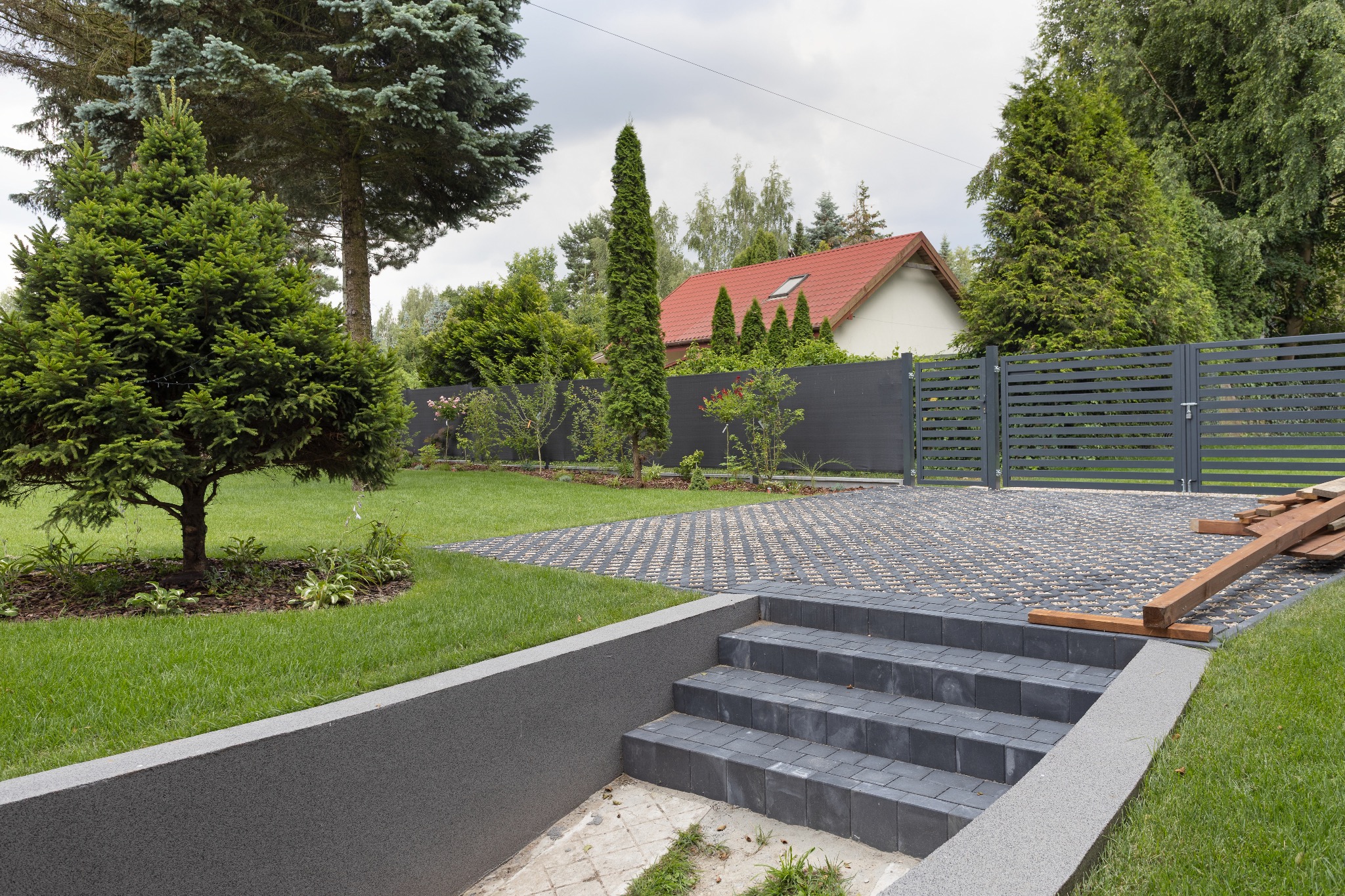 Nowoczesny podjazd z szarej kostki brukowej prowadzący do domu z czerwoną dachówką. Widoczne schody z kostki, trawnik i ogrodzenie panelowe w szarym kolorze.