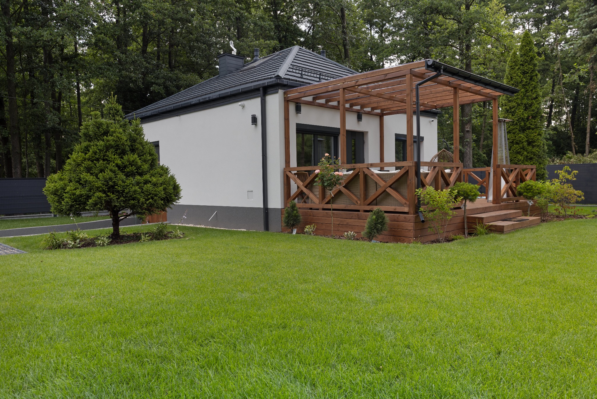 Biały dom z drewnianym tarasem i pergolą, otoczony zielonym trawnikiem i drzewami. Widoczny fragment ogrodzenia. Ujęcie z poziomu trawnika.