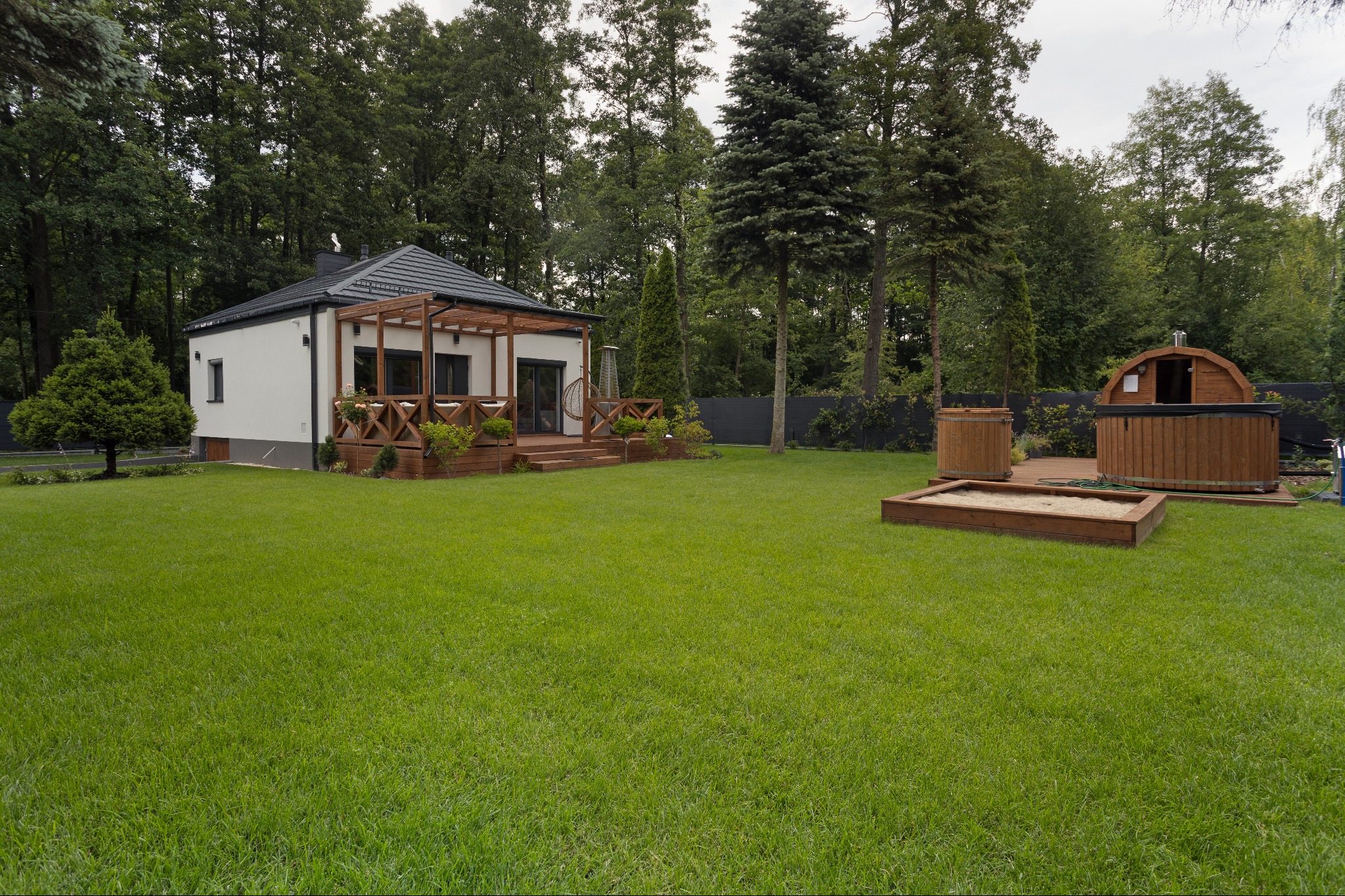 Nowoczesny dom z tarasem i drewnianą balią na zielonym trawniku, otoczony drzewami. Dom pomalowany na biało z szarym dachem.