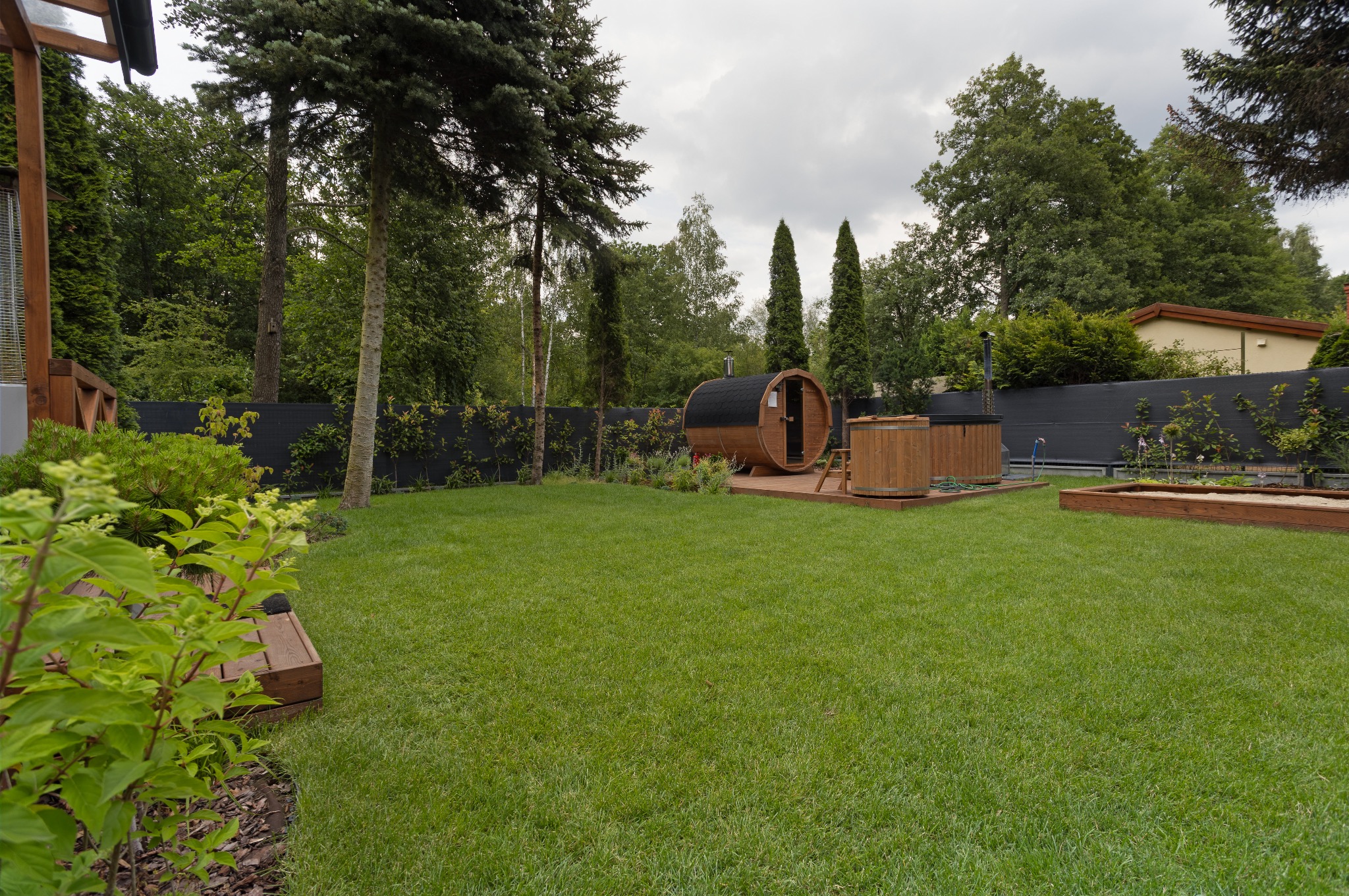 Nowoczesny ogród z drewnianą sauną beczką i jacuzzi na podeście. Zadbany trawnik, czarny płot i wysokie drzewa tworzą relaksującą przestrzeń.