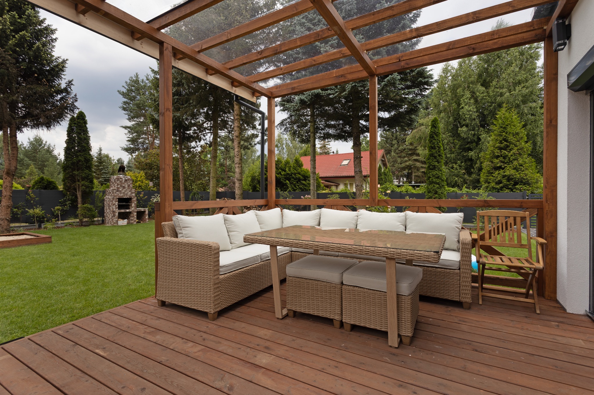 Drewniana pergola z meblami ogrodowymi z rattanu na tarasie. W tle zadbany trawnik i drzewa. Widoczny grill murowany.