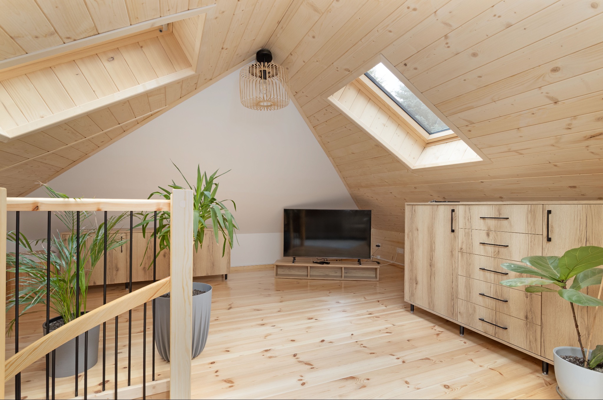 Jasne, drewniane poddasze ze skosami, oknem dachowym i meblami z jasnego drewna. Wnętrze doświetlone, z roślinami doniczkowymi i telewizorem.
