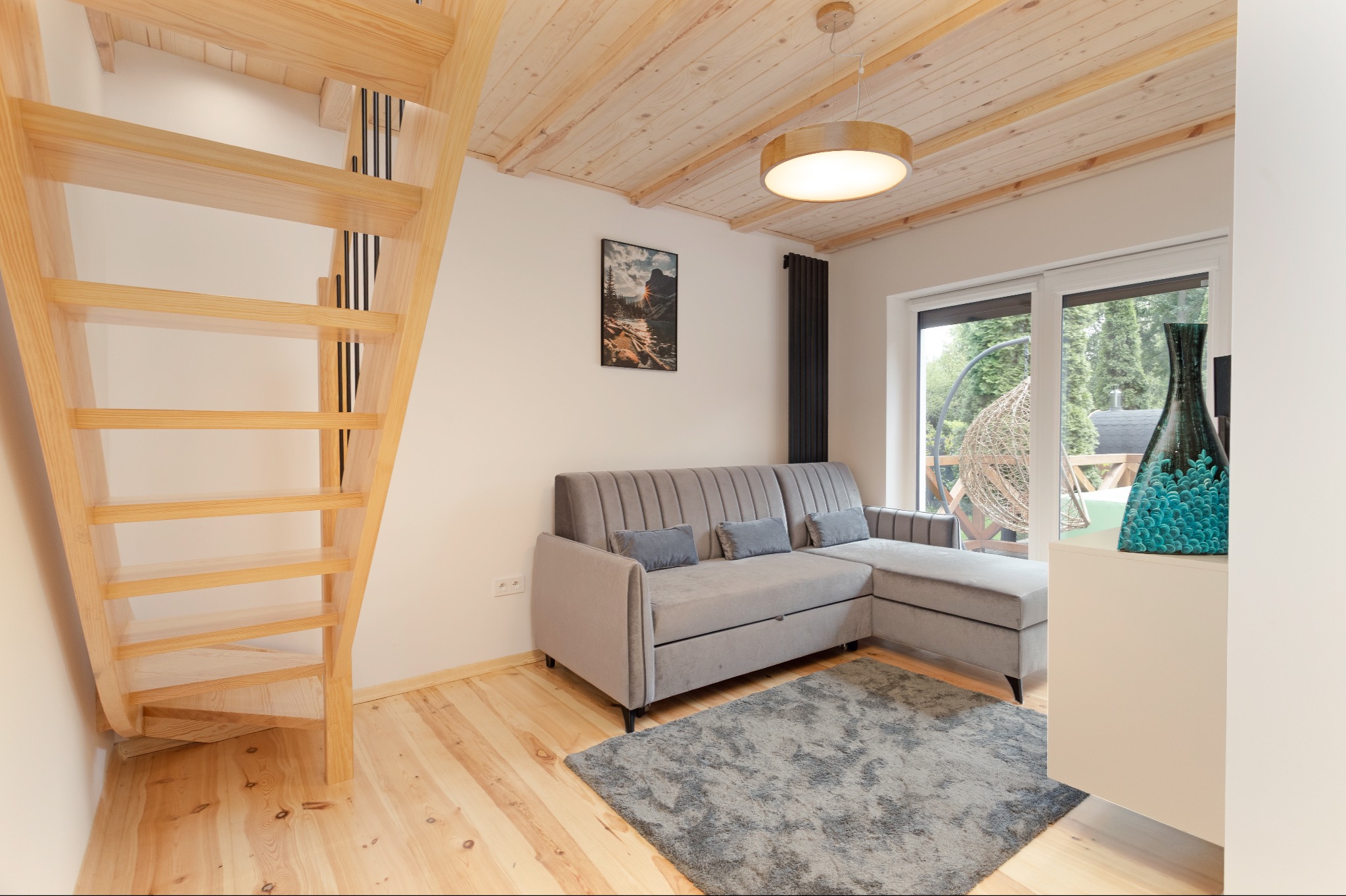 Jasne wnętrze pokoju z drewnianymi schodami, szarą sofą i ozdobną lampą. Widoczne drewniane wykończenie sufitu i podłogi, okno z widokiem na zewnątrz.