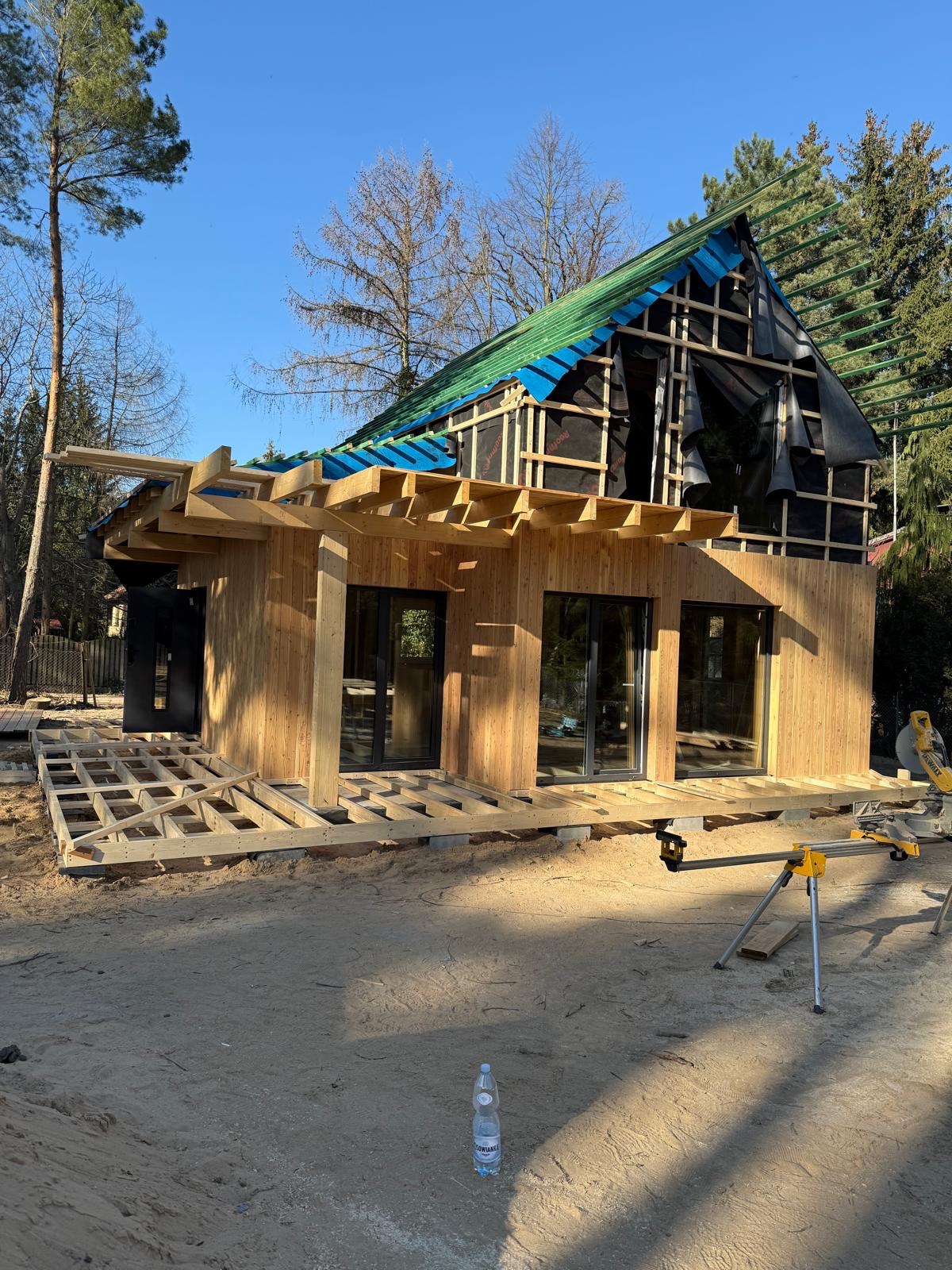 Budowa domu szkieletowego w trakcie: drewniana konstrukcja, okna wstawione, widoczne membrany dachowe i podkonstrukcja tarasu. Na pierwszym planie piasek i butelka wody.