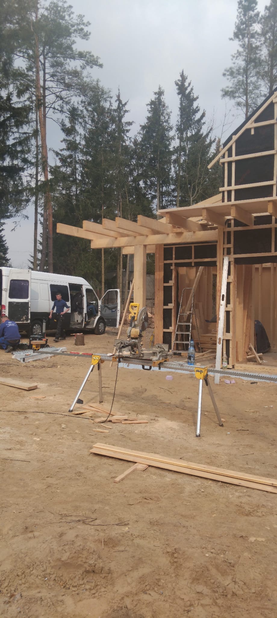 Budowa domu szkieletowego w lesie: widok na konstrukcję z drewna, narzędzia pracy (piła) i ekipę montażową przy białym busie. Etap wznoszenia ścian i zadaszenia.