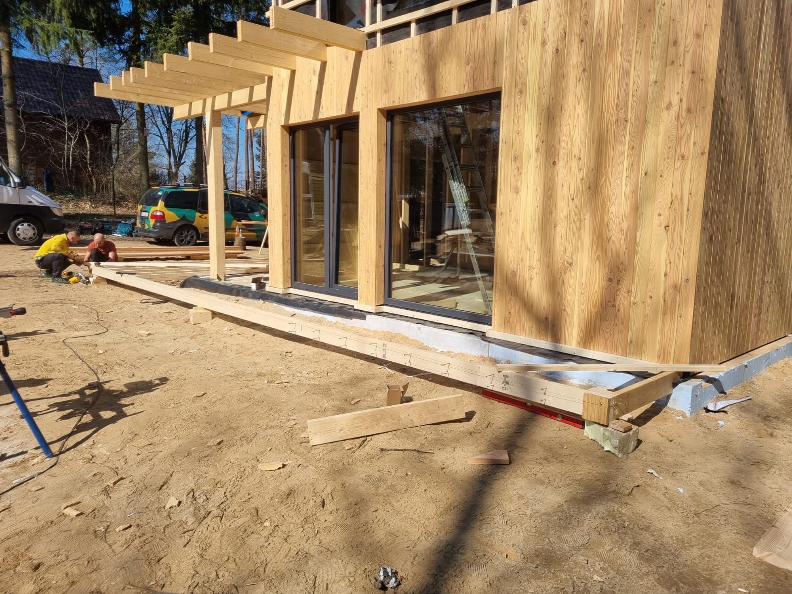 Budowa domu szkieletowego: montaż drewnianej elewacji i pergoli. Widoczne okna, fundament i pracujący robotnicy. Słoneczny dzień, naturalne oświetlenie.