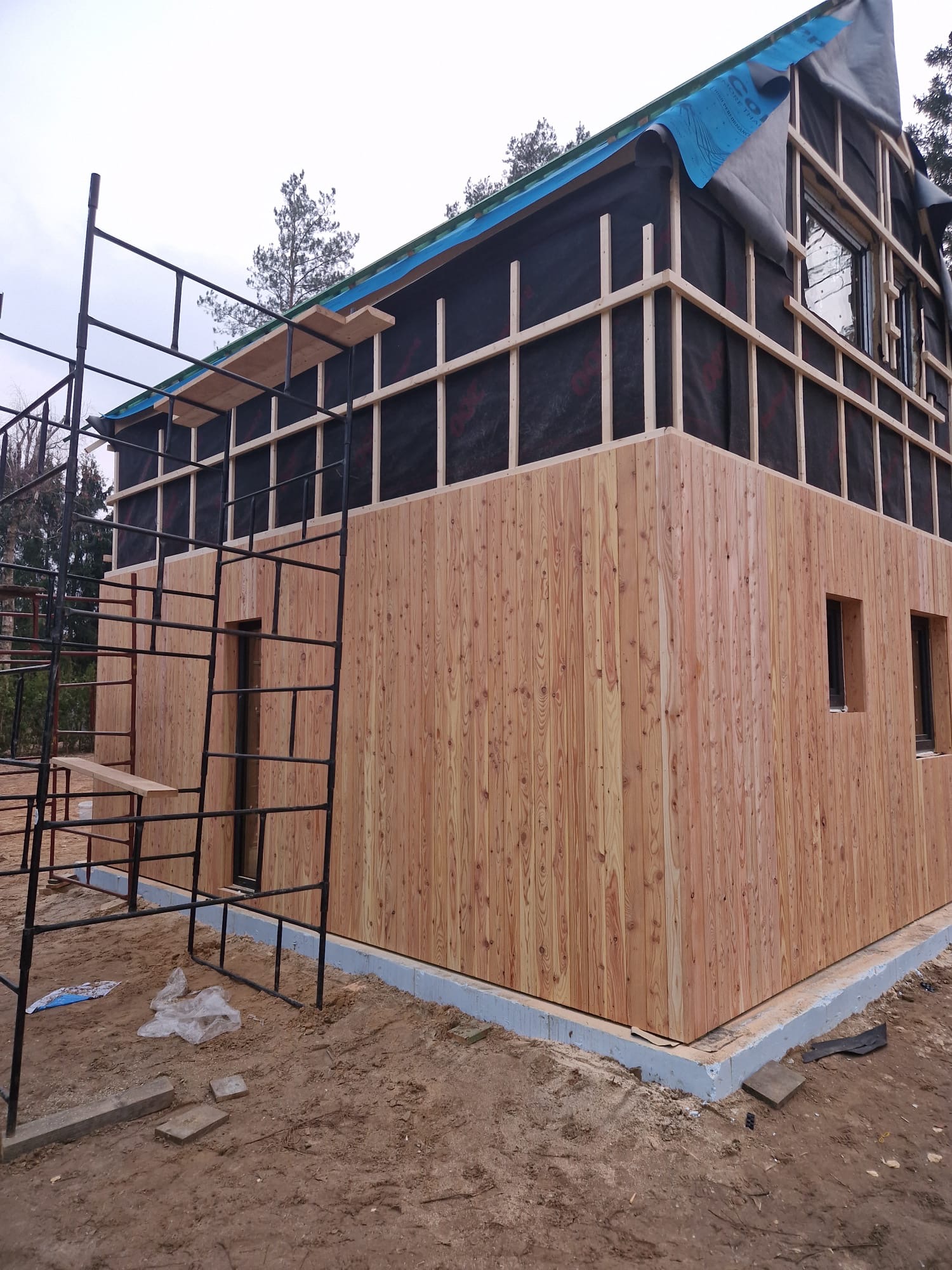 Dom w trakcie budowy: drewniana elewacja na parterze, czarna membrana na piętrze, rusztowania. Widoczne okna i fundamenty. Etap montażu elewacji.