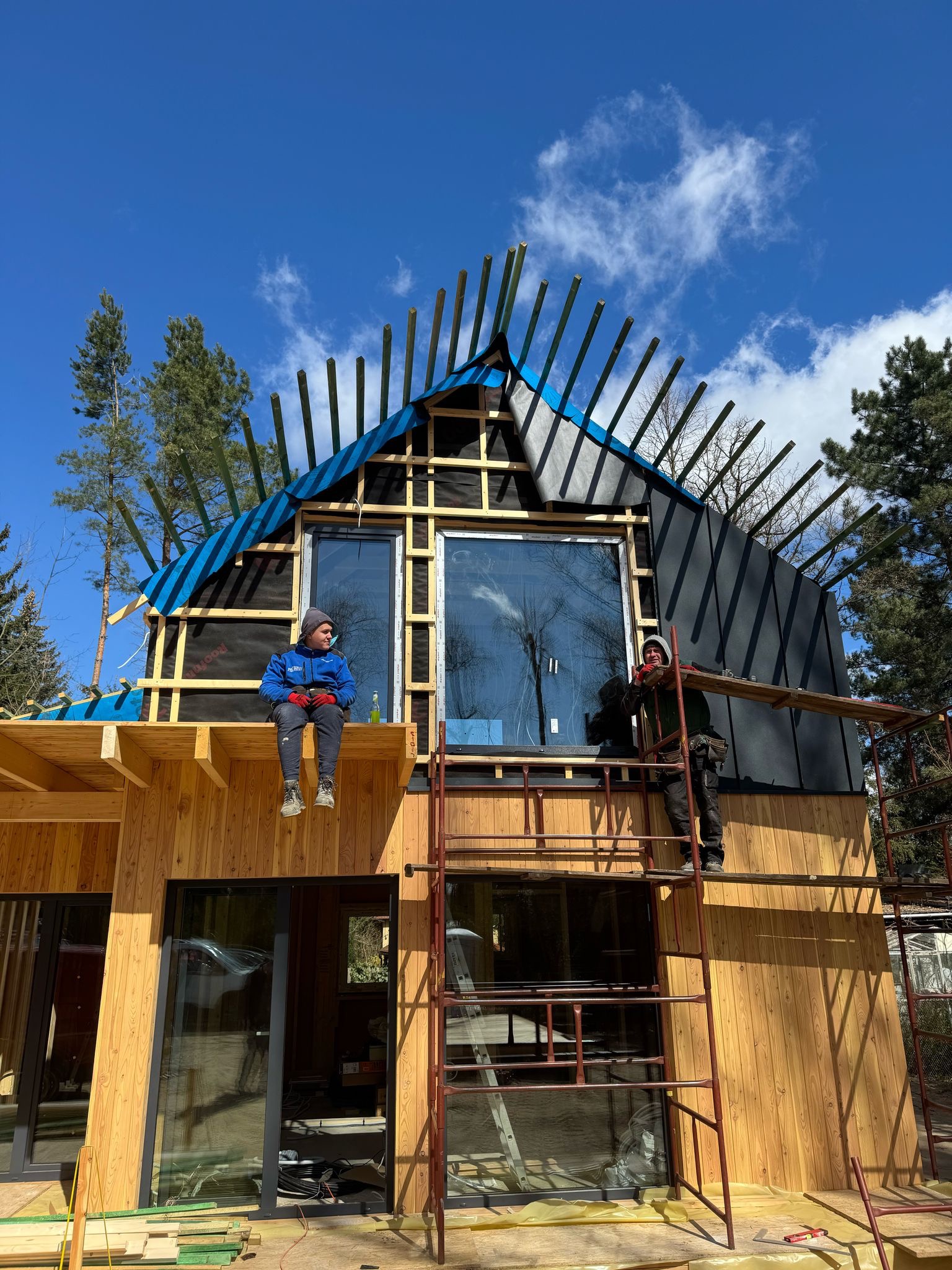 Budowa domu: dwaj robotnicy przy montażu drewnianej elewacji i dachu z membraną. Jeden siedzi na belce, drugi na rusztowaniu. Błękitne niebo w tle.