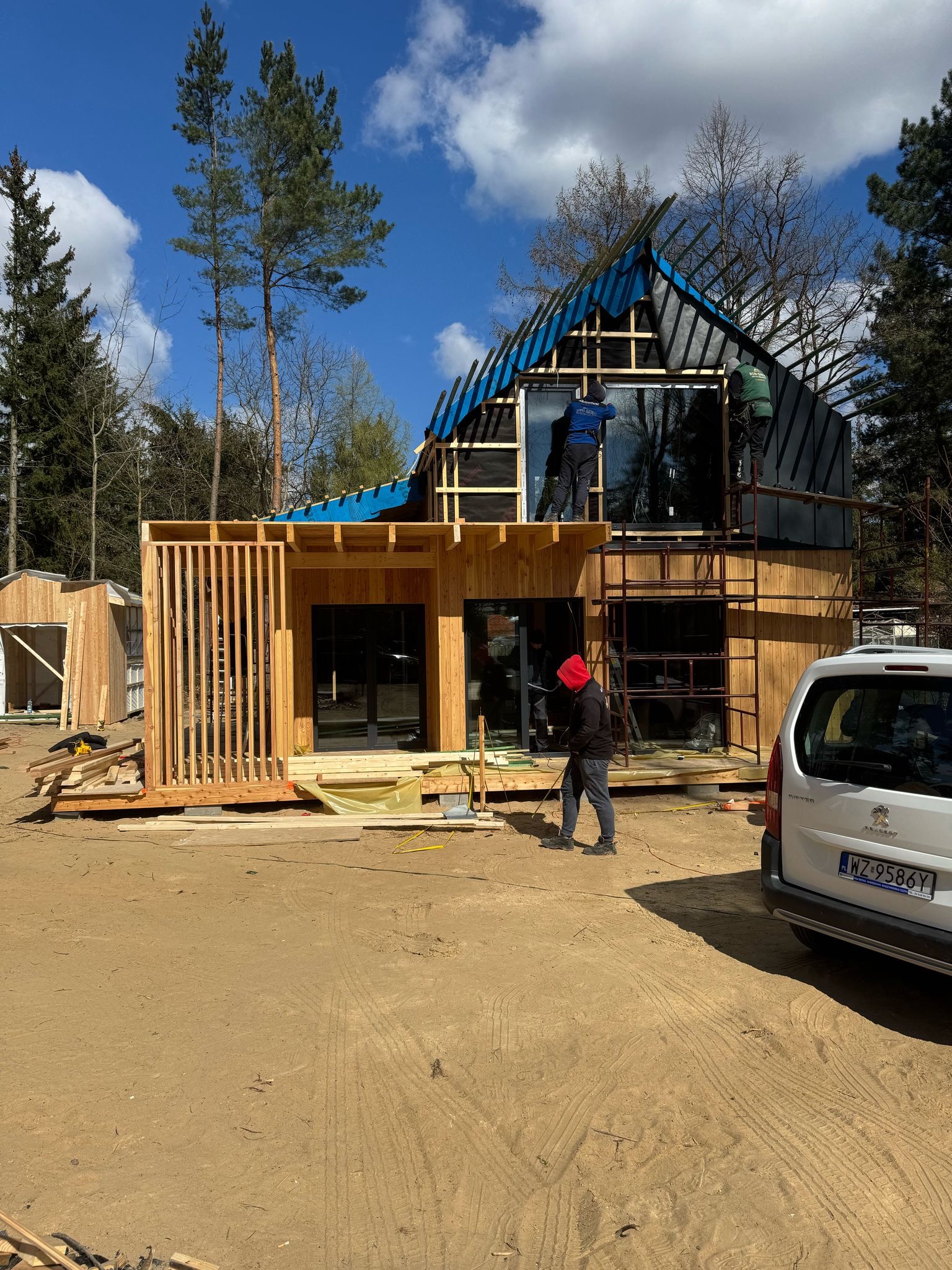 Budowa domu modułowego: drewniana konstrukcja, duże okna, robotnicy na rusztowaniu montują dach pokryty niebieską folią. Na pierwszym planie osoba w czerwonej bluzie.