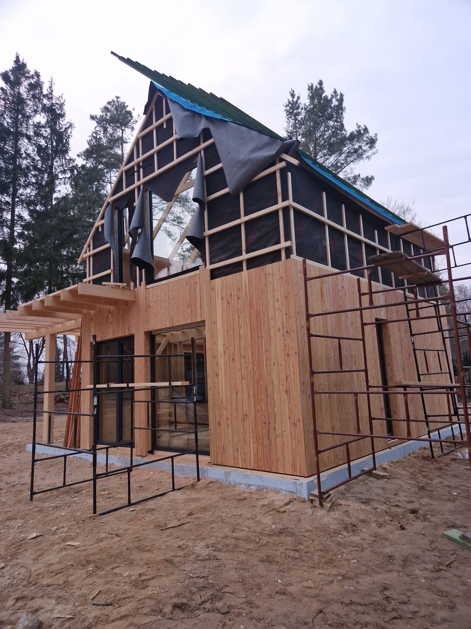 Drewniany dom w trakcie budowy, z widocznym szkieletem dachu i częściowo zamontowaną elewacją z desek. Na pierwszym planie rusztowania i teren budowy.