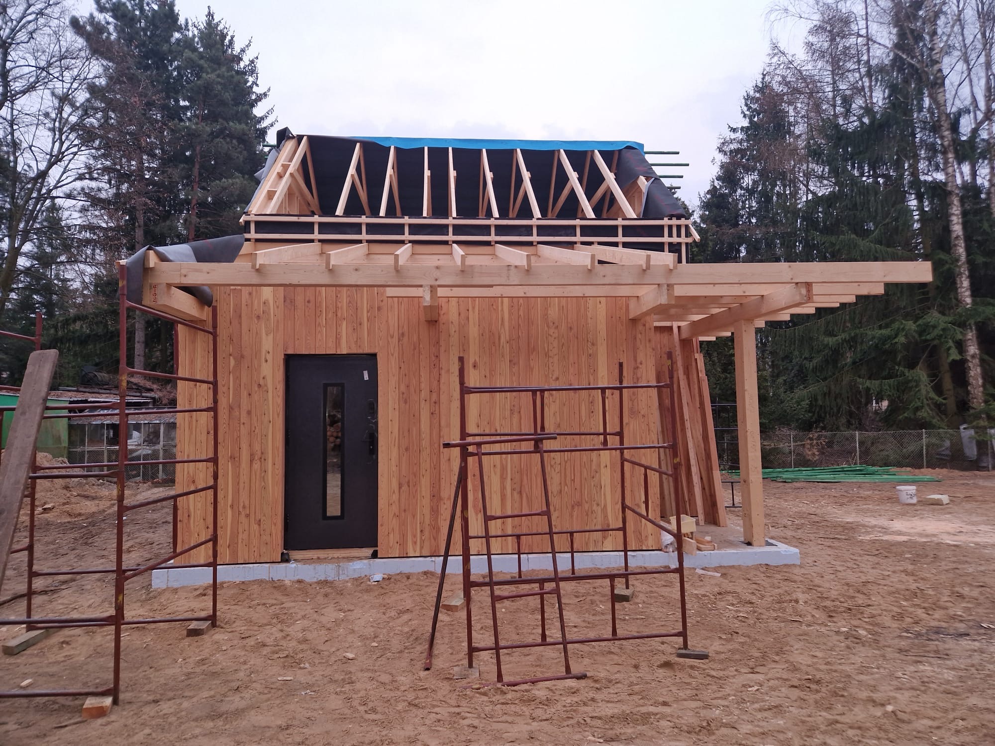 Budowa domu szkieletowego w trakcie: widoczne drewniane ściany, konstrukcja dachu z membraną, rusztowania. Dom z ciemnymi drzwiami, otoczony drzewami.