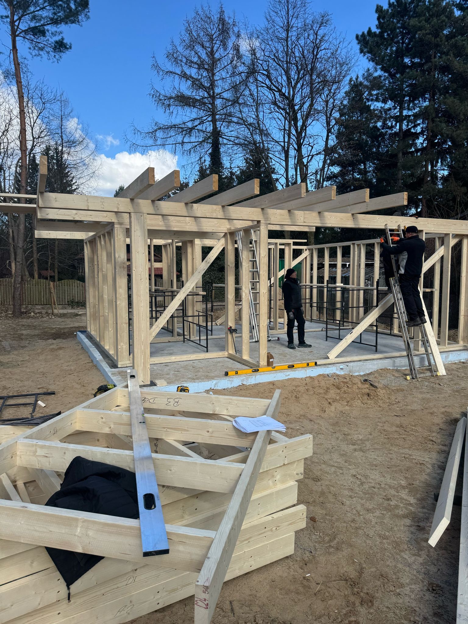 Budowa domu szkieletowego w toku: pracownicy montują drewnianą konstrukcję na fundamencie. Widoczne narzędzia i plany budowlane. Błękitne niebo w tle.