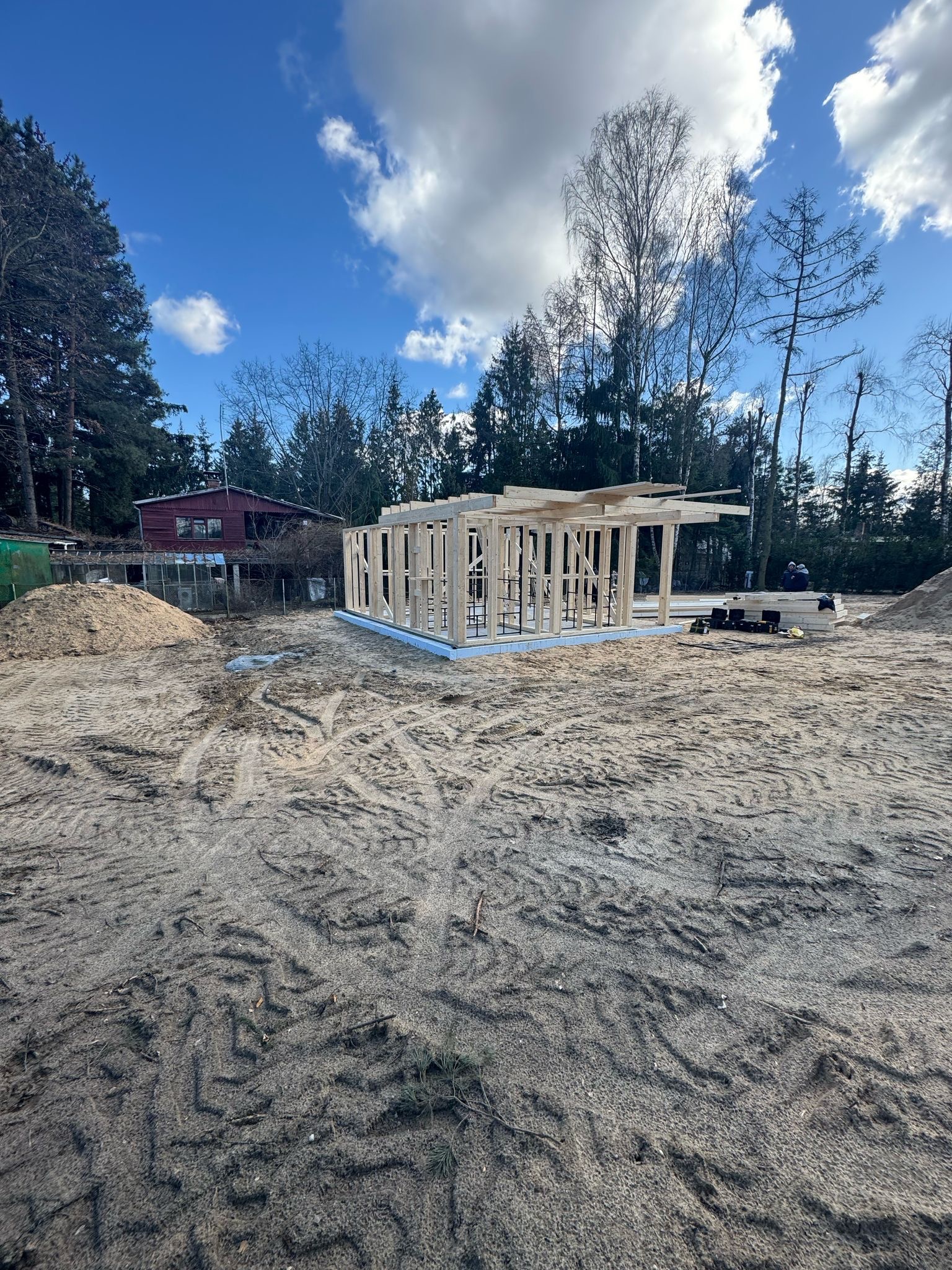 Wczesny etap budowy domu szkieletowego: drewniany szkielet na fundamencie, otoczony piaskiem i drzewami, pod błękitnym niebem z chmurami.
