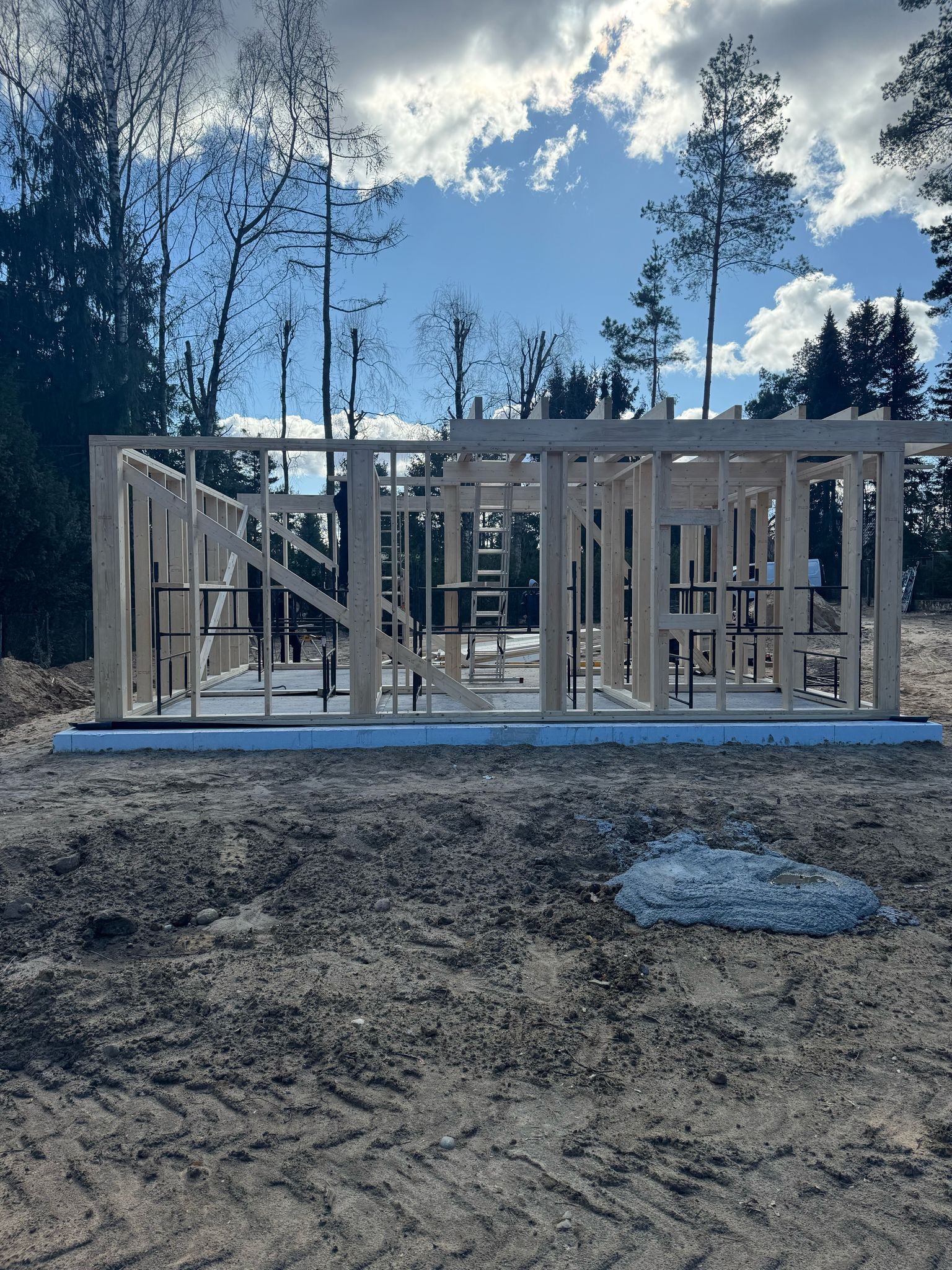 Surowa konstrukcja domu szkieletowego na fundamencie, widok z zewnątrz. Na pierwszym planie ziemia z odciśniętymi śladami opon, w tle drzewa i błękitne niebo z chmurami.