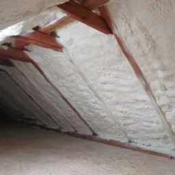 Poddasze z drewnianą konstrukcją pokryte grubą warstwą białej pianki izolacyjnej. Widoczna tekstura pianki i fragment podłogi.