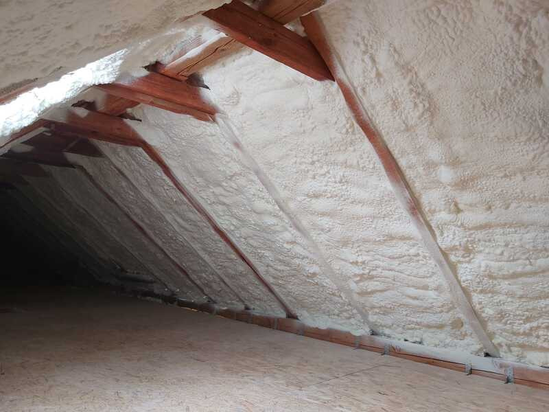 Poddasze z drewnianą konstrukcją pokryte grubą warstwą białej pianki izolacyjnej. Widoczna tekstura pianki i fragment podłogi.