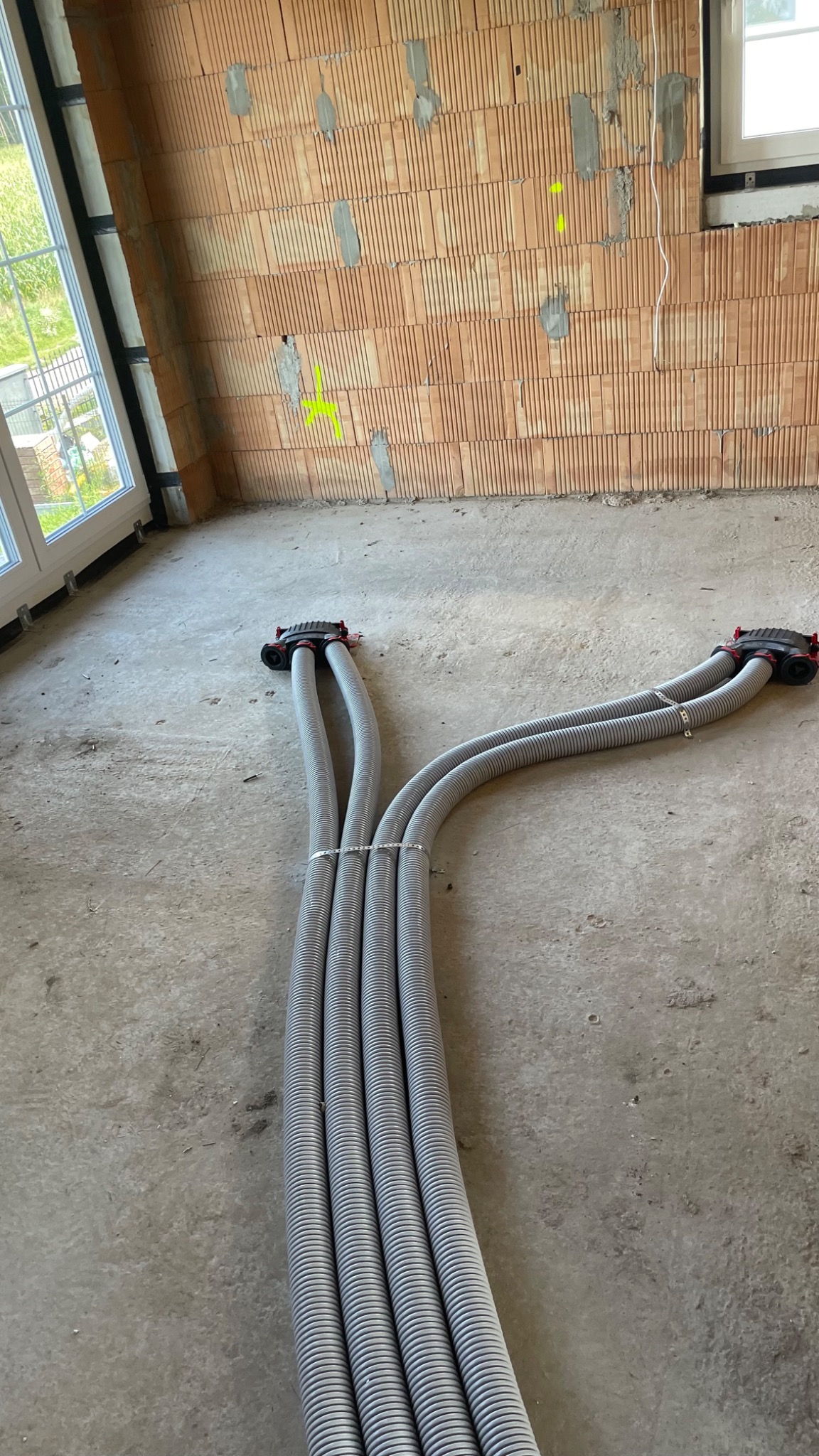 Szare, karbowane rury wentylacyjne w surowym wnętrzu z ceglaną ścianą i oknem. Etap instalacji rekuperacji w nowym budynku. Elementy montażowe widoczne.