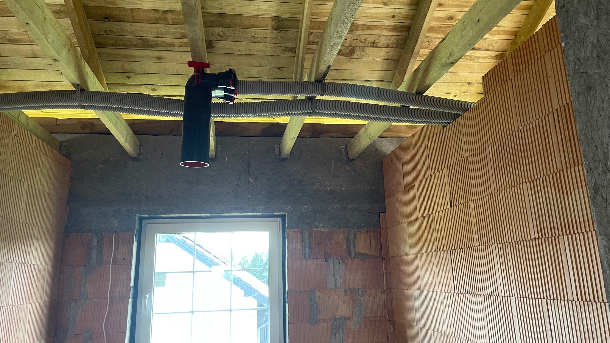 Wnętrze w trakcie instalacji rekuperacji: widoczne rury wentylacyjne pod drewnianym stropem i centrala rekuperacyjna, ściany z cegły dziurawki.