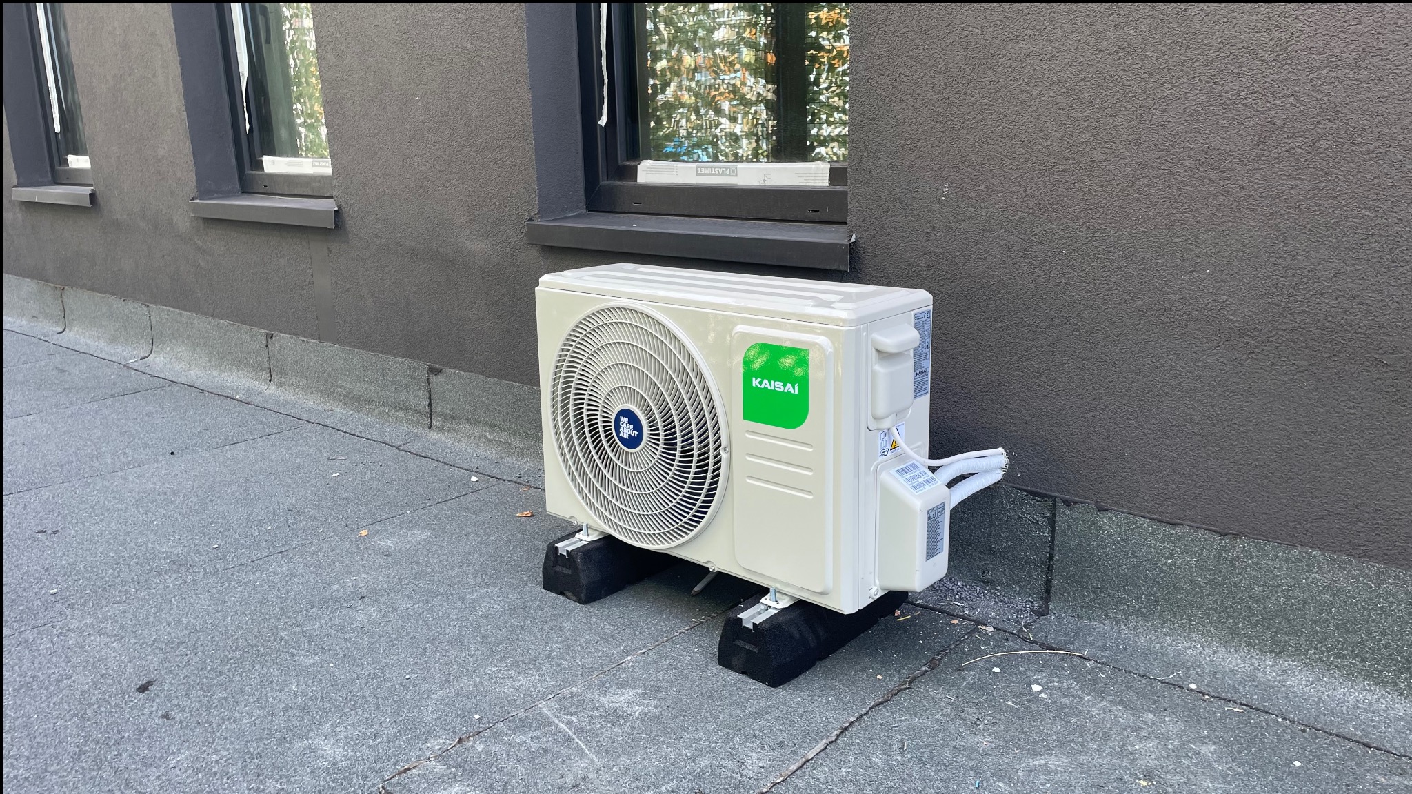 Biały klimatyzator Kaisai, zamontowany na zewnątrz budynku z szarą elewacją, widoczny fragment okna. Ujęcie z poziomu podłoża.