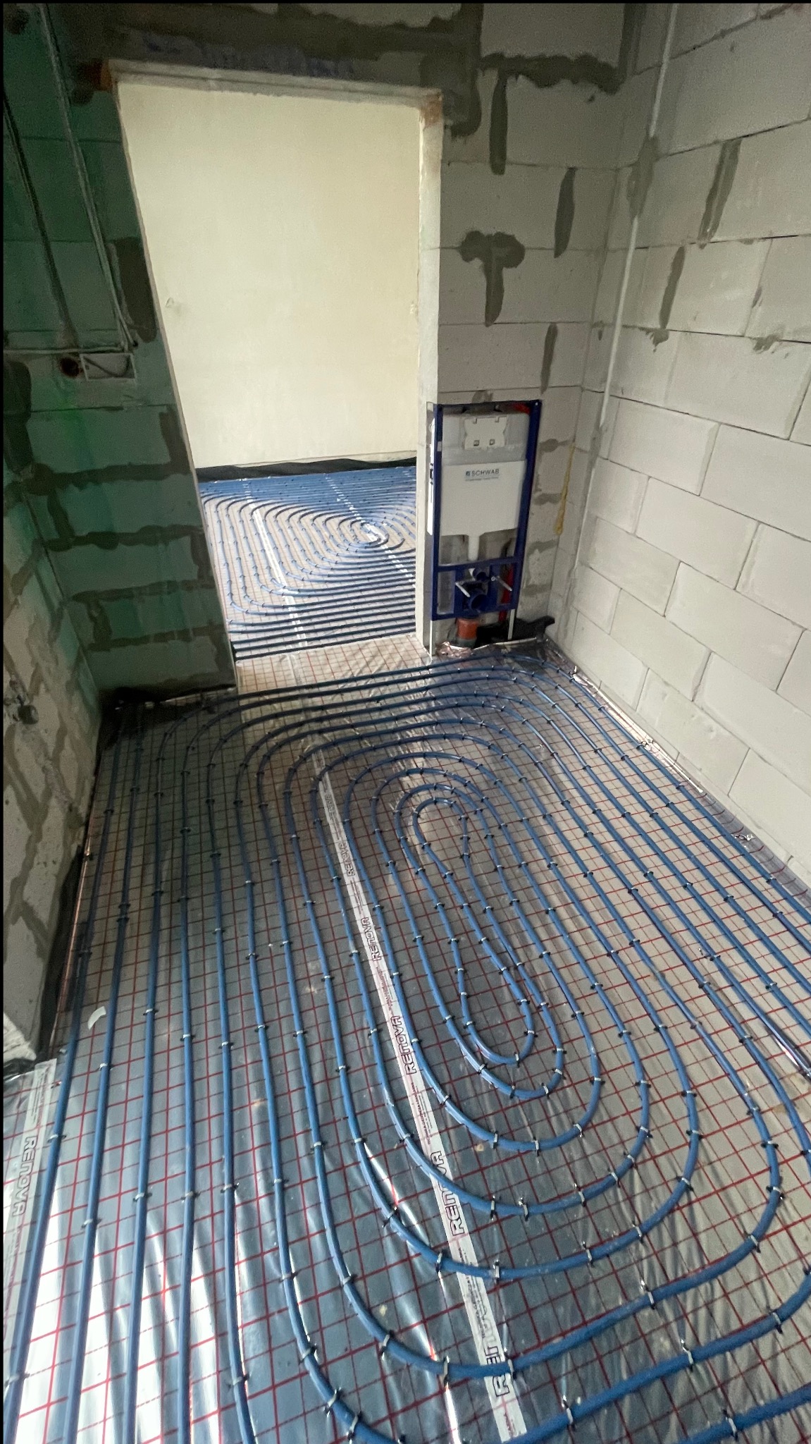 Niebieskie rury ogrzewania podłogowego ułożone na folii z siatką w surowym wnętrzu. Widoczna instalacja podtynkowa WC. Etap prac przed wylewką.