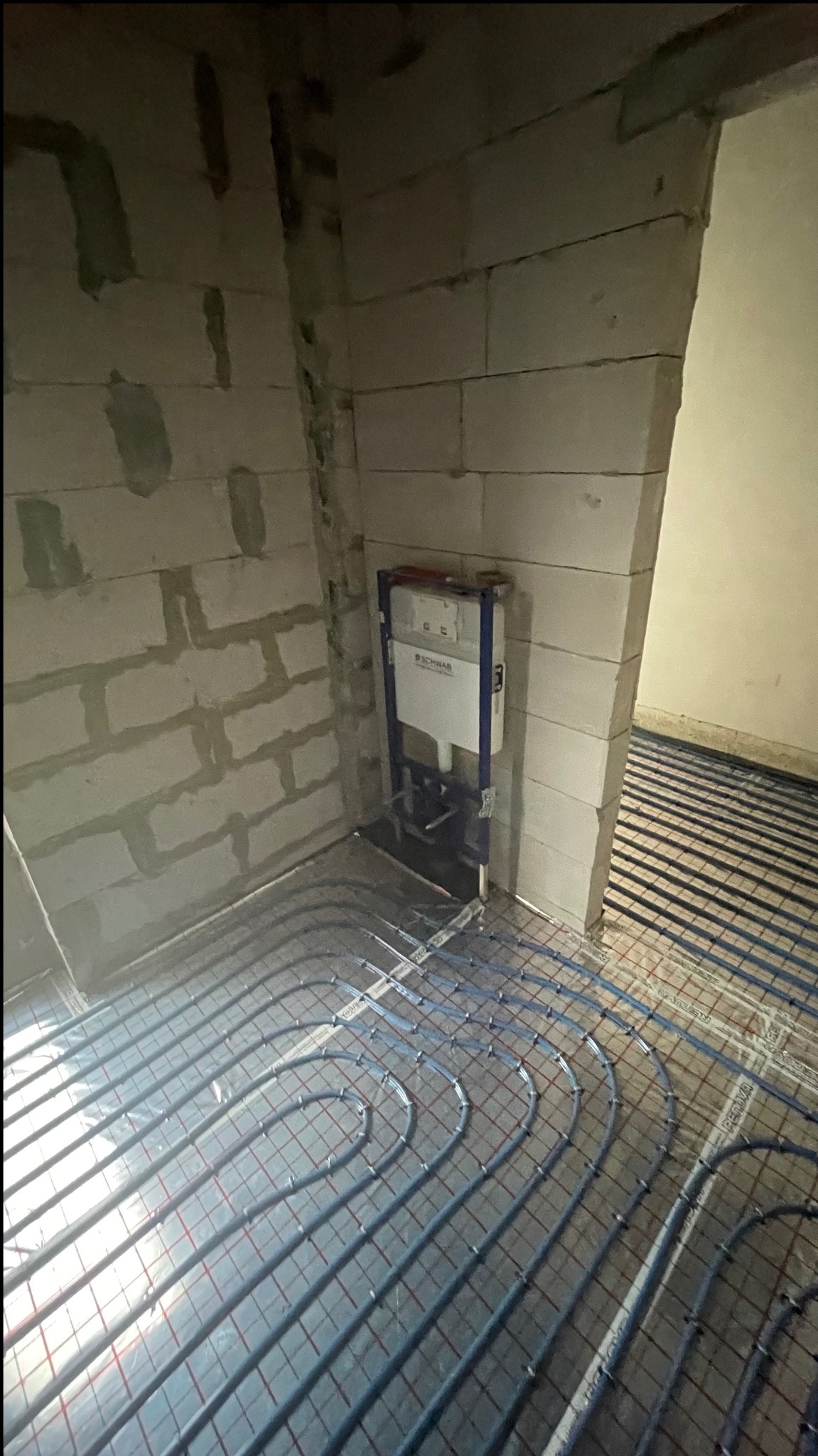 Instalacja ogrzewania podłogowego z zamontowanym stelażem podtynkowym WC marki Schwab, widoczne rury w szarej folii na podłodze w surowym pomieszczeniu.