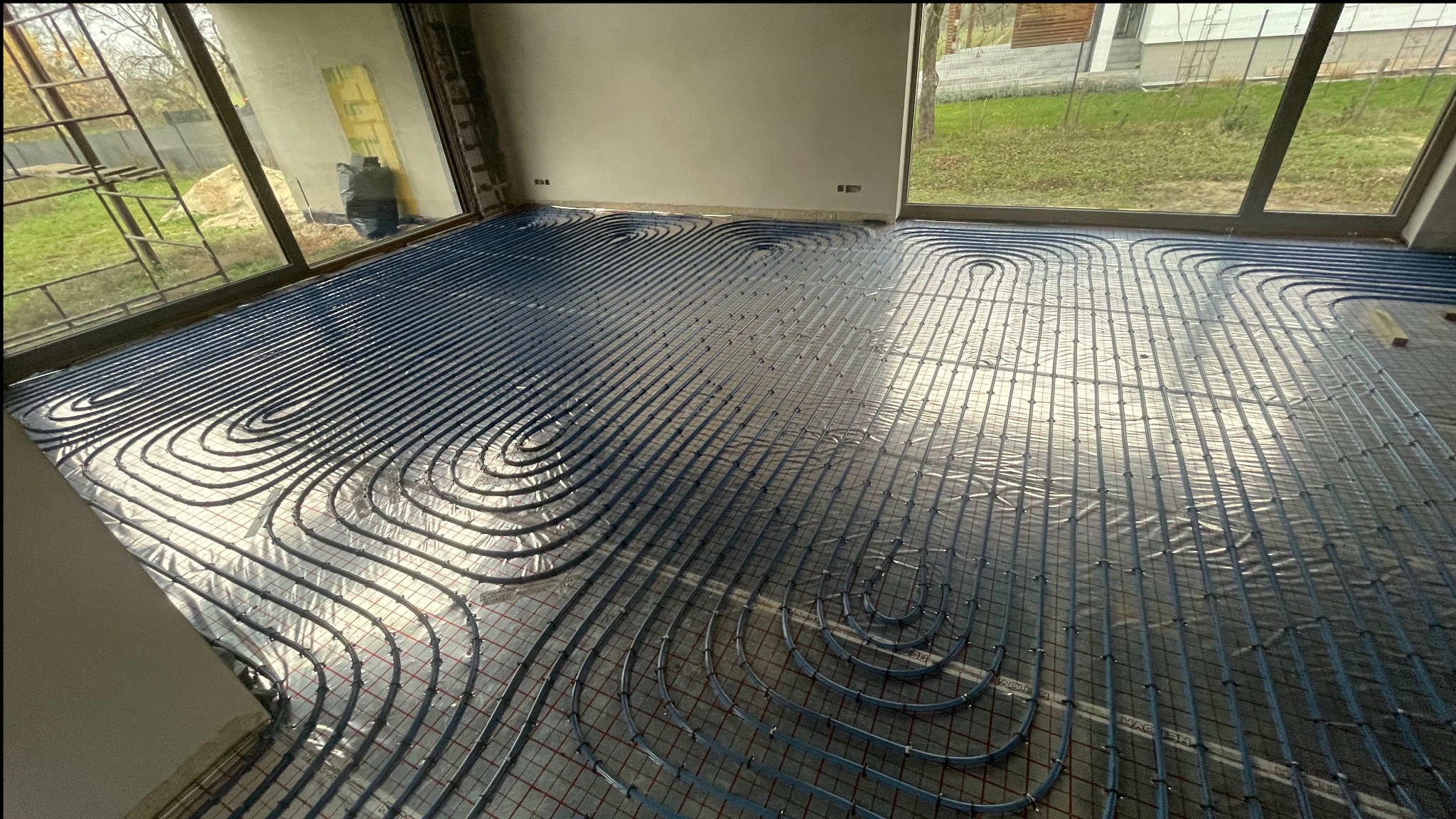 Ułożone rury ogrzewania podłogowego na folii izolacyjnej w surowym wnętrzu z dużymi oknami. Widoczne pętle rur i siatka montażowa. Budynek w tle.