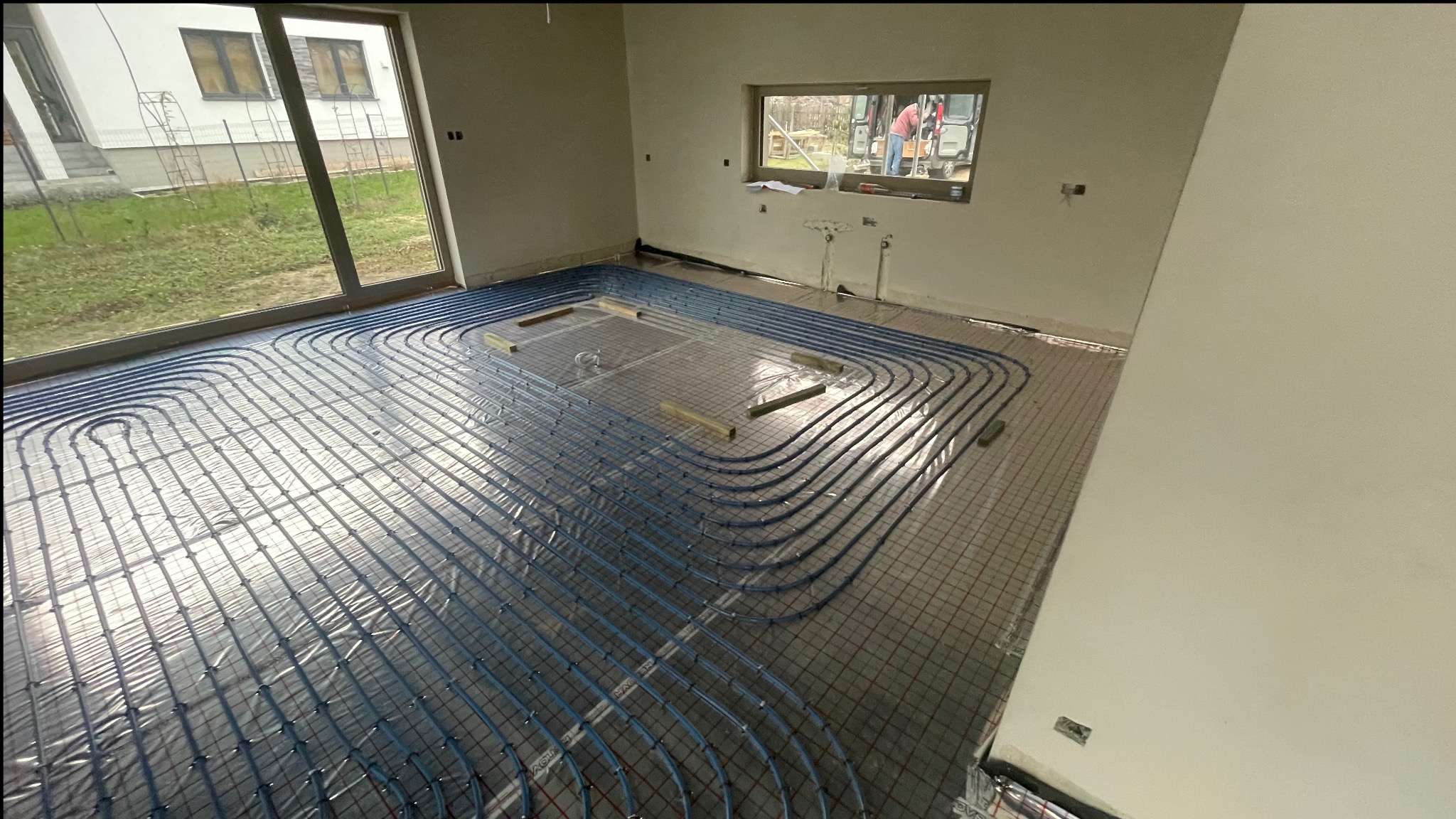 Niebieskie rury ogrzewania podłogowego rozłożone na siatce w surowym wnętrzu. Widoczne okno z widokiem na zewnątrz i elementy instalacji na ścianie.