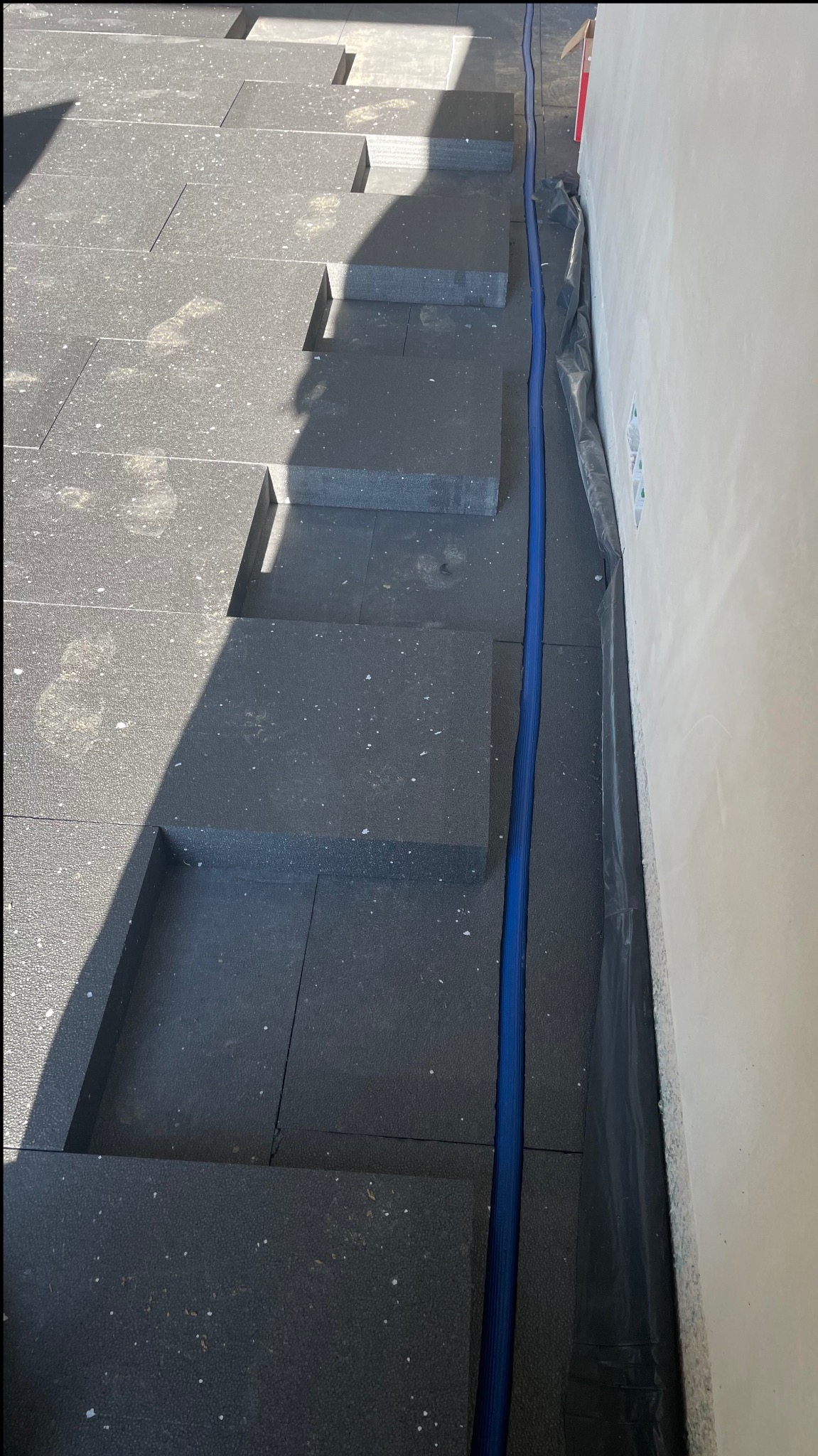 Przygotowanie podłoża pod ogrzewanie podłogowe: rozłożony styropian EPS z wycięciami na rury, niebieska rura wzdłuż ściany, folia ochronna.