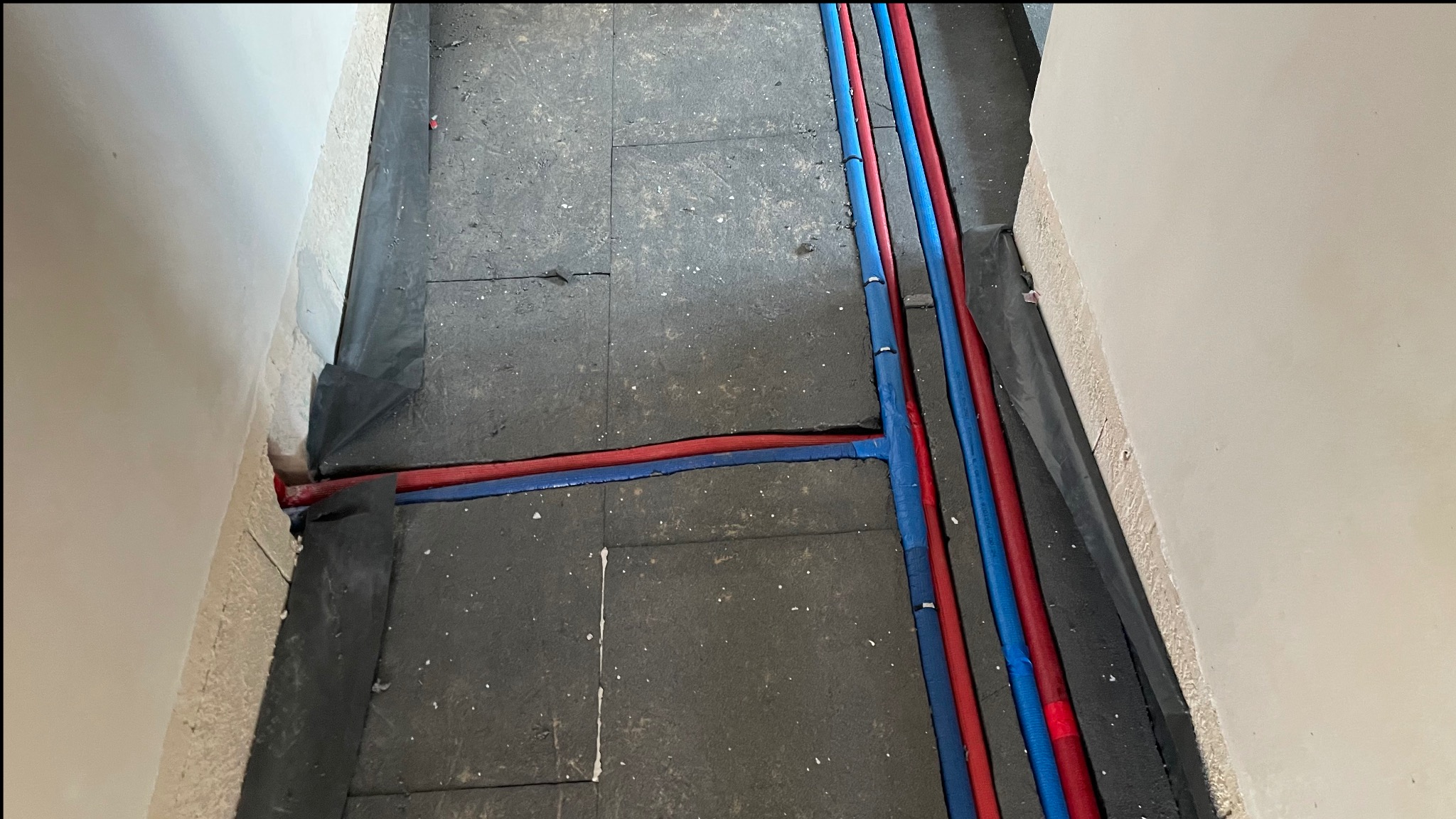 Instalacja rur grzewczych (czerwone i niebieskie) na podłożu. Prace hydrauliczne w Toruniu przed wylaniem posadzki. Widok z góry, rury ułożone wzdłuż ściany.