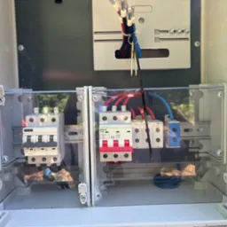 Rozdzielnica elektryczna w obudowie z widocznymi bezpiecznikami, przewodami i elementami montażowymi. Widok z bliska na wnętrze skrzynki elektrycznej.