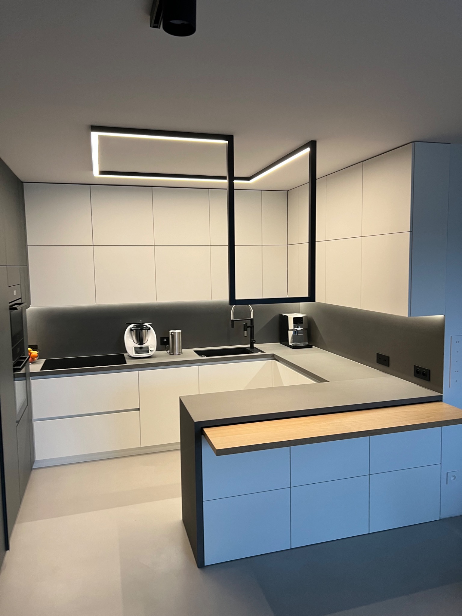 Nowoczesna kuchnia z szarym blatem, białymi szafkami i oryginalnym oświetleniem. Minimalistyczny design, czarne akcenty, sprzęt AGD na blacie.