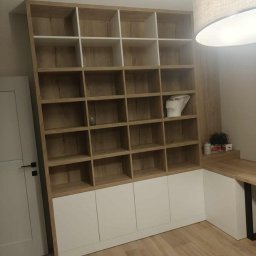 Magdalena Oniszczuk - Regał na wymiar z białymi frontami i drewnianymi półkami, część aranżacji biurowej. Widoczny fragment blatu biurka i lampy. Ujęcie z dołu, podkreślające wysokość mebla.