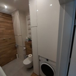 Magdalena Oniszczuk - Biała szafa w łazience z wbudowaną pralką, widok na toaletę i drewnianą ścianę w tle. Funkcjonalna zabudowa w małej przestrzeni.