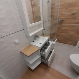 Magdalena Oniszczuk - Nowoczesna biała szafka łazienkowa z otwartymi półkami i wysuwanymi szufladami, z umywalką i lustrem na tle kabiny prysznicowej z drewnopodobnymi płytkami.