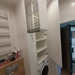 Magdalena Oniszczuk - Biała szafa z koszem cargo i półkami, zabudowana nad pralką w łazience. Widoczny fragment blatu i akcesoria montażowe. Otwarta szafka.