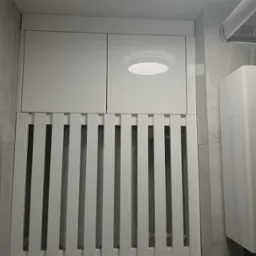 Biała szafka na wymiar z żaluzjowym frontem, zakrywająca grzejnik w łazience. Minimalistyczny design, szare płytki w tle i okrągłe oświetlenie.