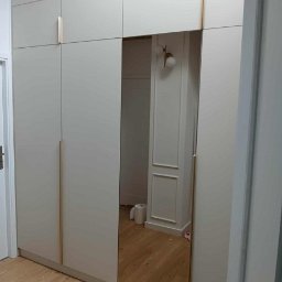 Magdalena Oniszczuk - Jasna szafa wnękowa z lustrem, złotymi uchwytami i drewnianą nadstawką, wkomponowana w jasne wnętrze. Widoczne detale wykończenia ścian.
