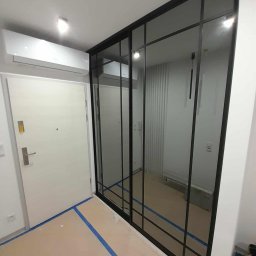 Magdalena Oniszczuk - Nowoczesna szafa wnękowa z czarnymi ramami i lustrzanymi drzwiami przesuwnymi, widoczna w korytarzu z zabezpieczoną podłogą i klimatyzacją.