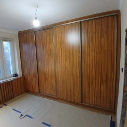 Magdalena Oniszczuk - Drewniana szafa wnękowa z przesuwnymi drzwiami, widoczna w pokoju z zabezpieczoną podłogą. Ujęcie z perspektywy podłogi, z widocznym oknem po lewej stronie.