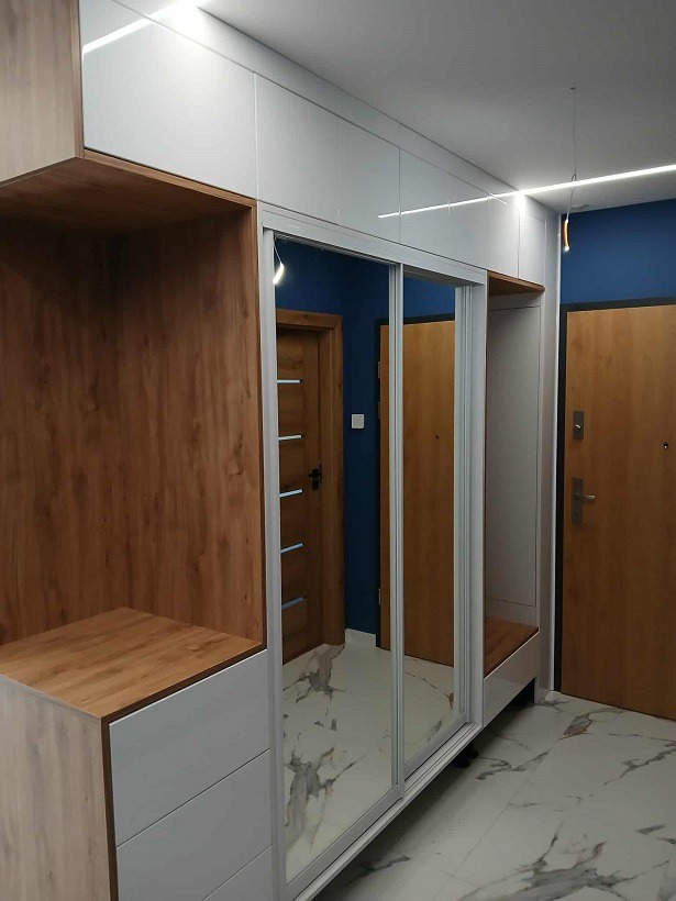 Nowoczesna szafa wnękowa z przesuwnymi drzwiami lustrzanymi i elementami drewnopodobnymi, wkomponowana w korytarz z marmurową podłogą i niebieską ścianą.