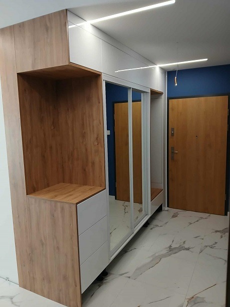 Szafa wnękowa z lustrzanymi drzwiami przesuwnymi, drewnopodobne wykończenie i białe fronty szuflad, oświetlenie LED w suficie, marmurowa podłoga, widok na drzwi.