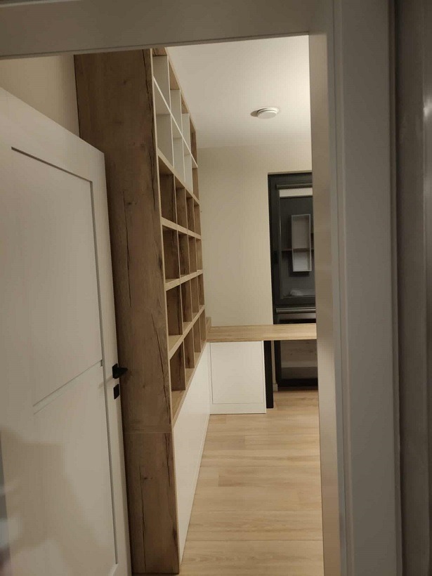 Biała szafa wnękowa z drewnianymi półkami, widoczna zza uchylonych drzwi. Minimalistyczny design, jasne wnętrze, podłoga z paneli.