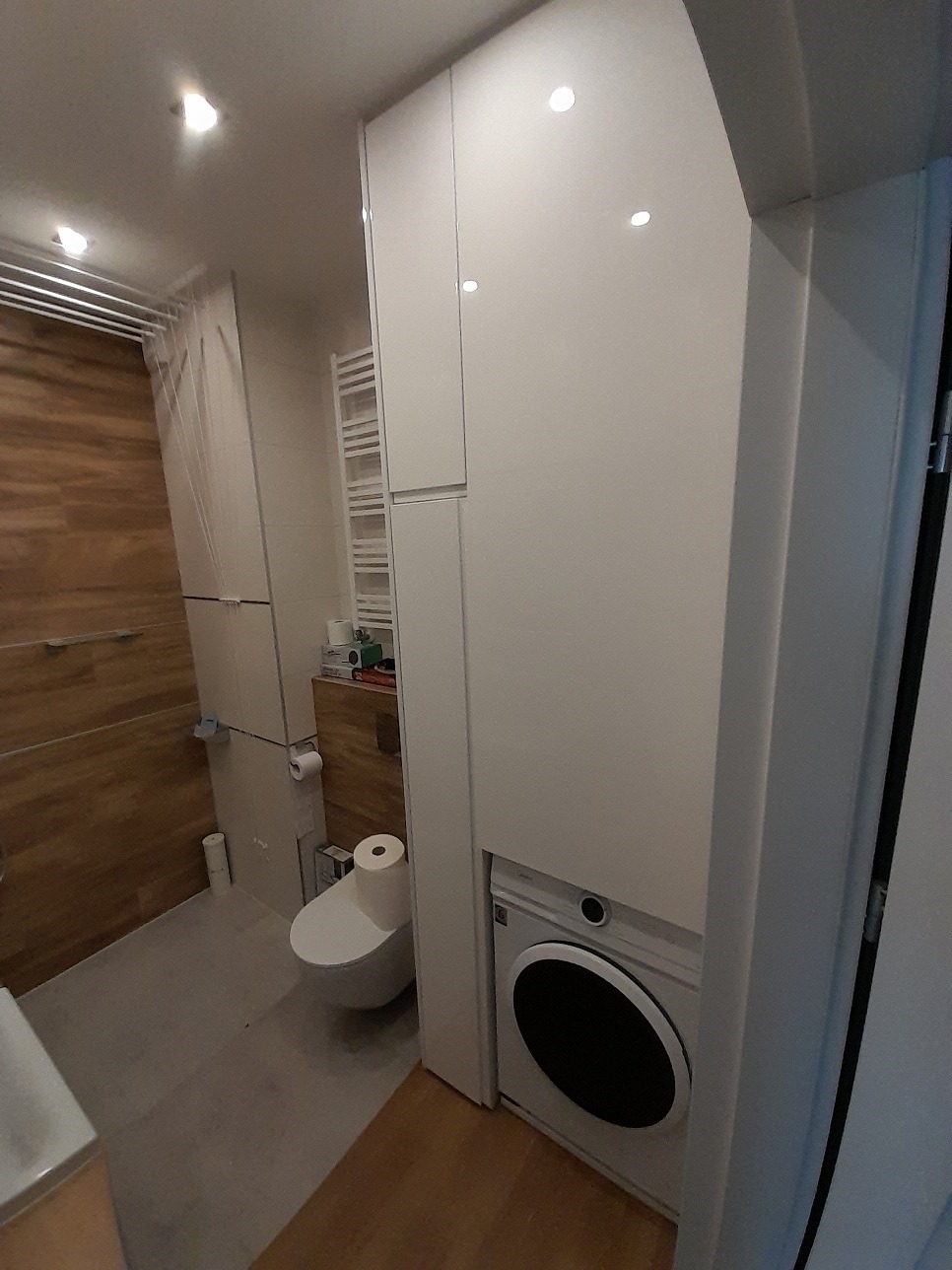 Biała szafa w łazience z wbudowaną pralką, widok na toaletę i drewnianą ścianę w tle. Funkcjonalna zabudowa w małej przestrzeni.