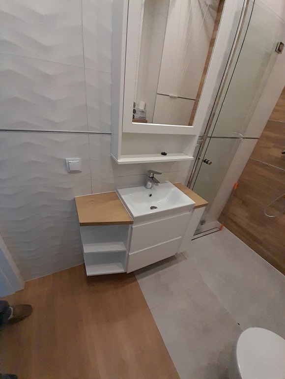 Biała szafka łazienkowa z umywalką i lustrem, drewniany blat po lewej stronie, Lublin. Nowoczesny design, płytki 3D na ścianie, kabina prysznicowa w tle.