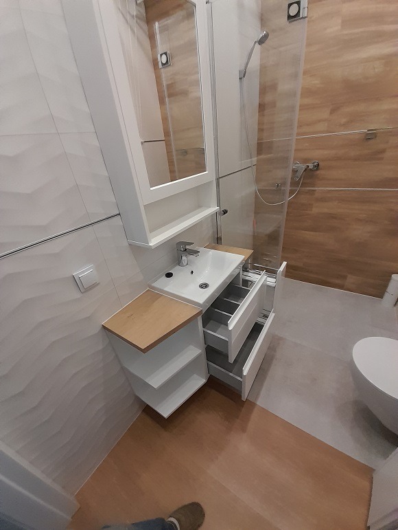 Nowoczesna biała szafka łazienkowa z otwartymi półkami i wysuwanymi szufladami, z umywalką i lustrem na tle kabiny prysznicowej z drewnopodobnymi płytkami.