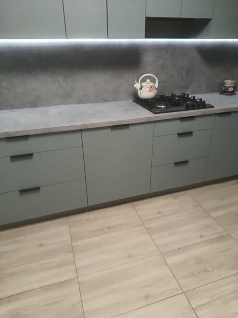 Jasnoszara kuchnia z blatem imitującym beton, oświetleniem LED pod szafkami i czajnikiem na płycie grzewczej. Minimalistyczny design.