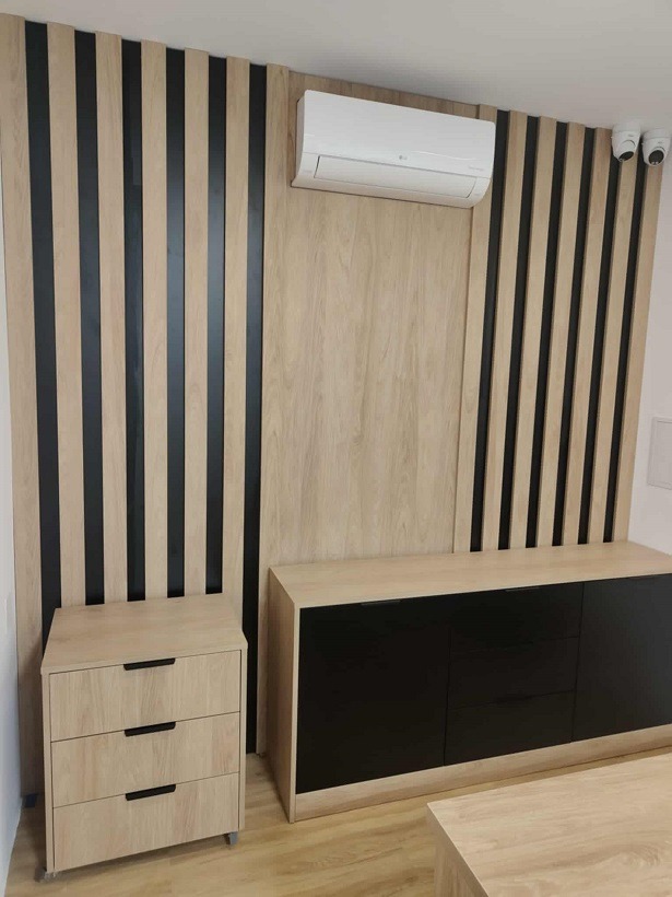 Nowoczesna komoda z czarnymi frontami i szafka nocna z trzema szufladami w pokoju z dekoracyjną ścianą z drewnianych paneli z klimatyzatorem i kamerami.
