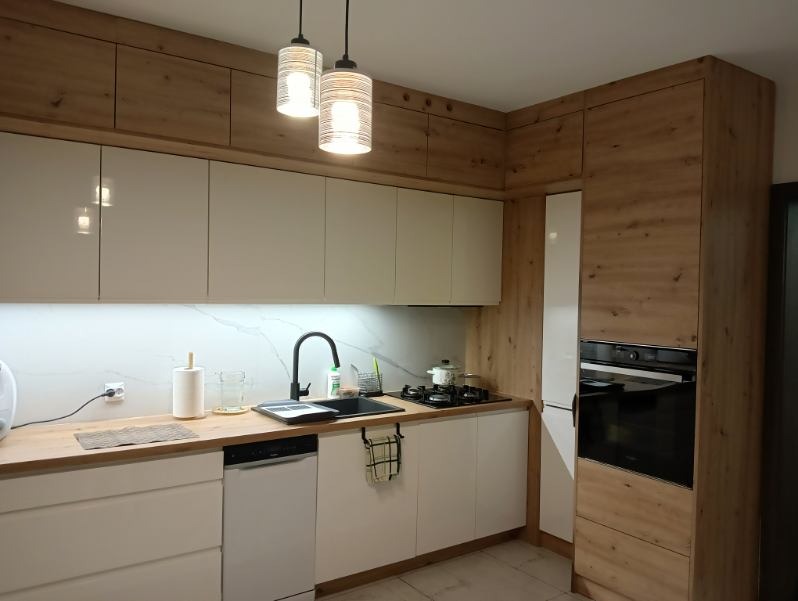 Nowoczesna kuchnia z białymi frontami i drewnianymi akcentami. Widoczne wiszące lampy, czarny zlew i sprzęt AGD. Blat drewniany.