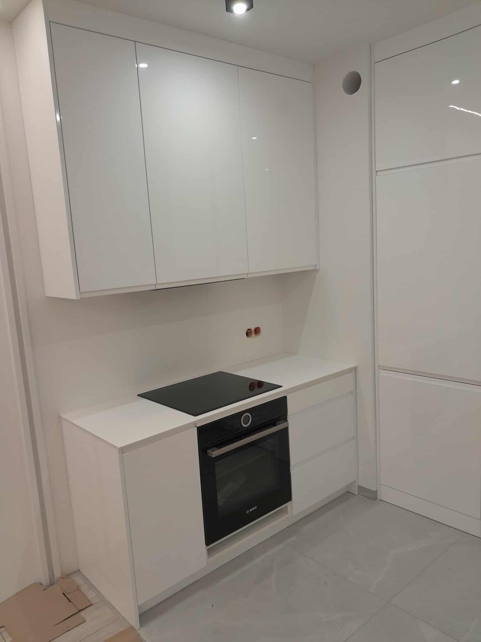 Biała, minimalistyczna kuchnia z błyszczącymi frontami szafek, czarną płytą indukcyjną i piekarnikiem Bosch. Widoczne gniazdka nad blatem i szare płytki podłogowe.